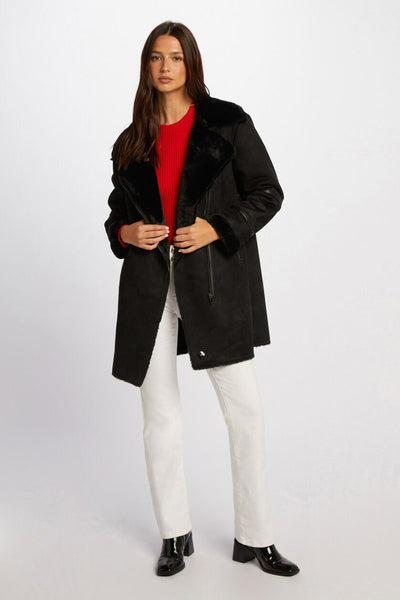 Veste longue noire de Morgan de Toi avec col en fourrure portée sur un pull rouge et un jean blanc.