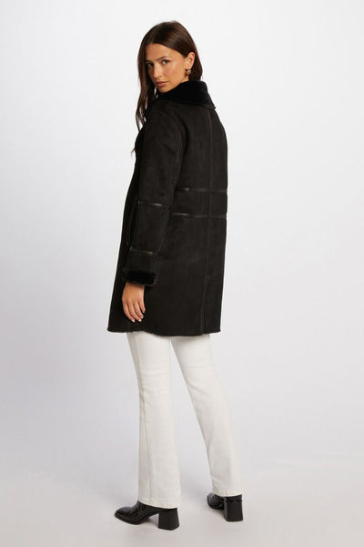 Veste longue noire par Morgan de Toi avec col en fausse fourrure, portée avec un jean blanc et des bottes noires.