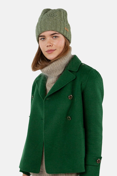 Bonnet vert en maille torsadée de BARTS, porté avec un manteau vert et un pull à col roulé beige.
