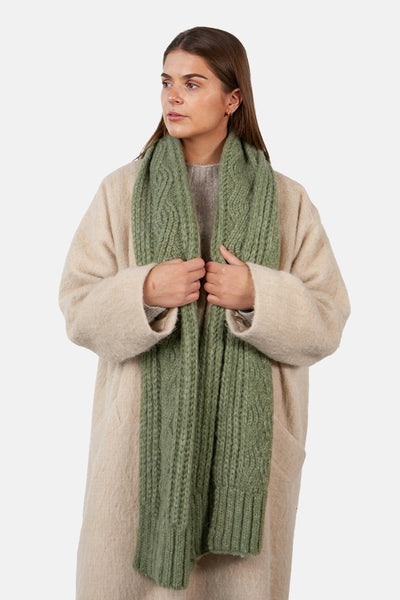 Écharpe verte par BARTS, à motif torsadé, portée avec un manteau beige texturé.
