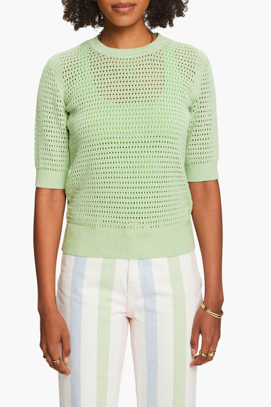 Pull à col rond en maille verte d'Esprit, porté avec un pantalon rayé multicolore et des bijoux dorés.
