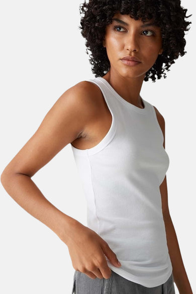 Top blanc sans manches de OPUS à côtes, porté par une femme aux cheveux bouclés, avec un léger poil sous les bras, et un pantalon gris.
