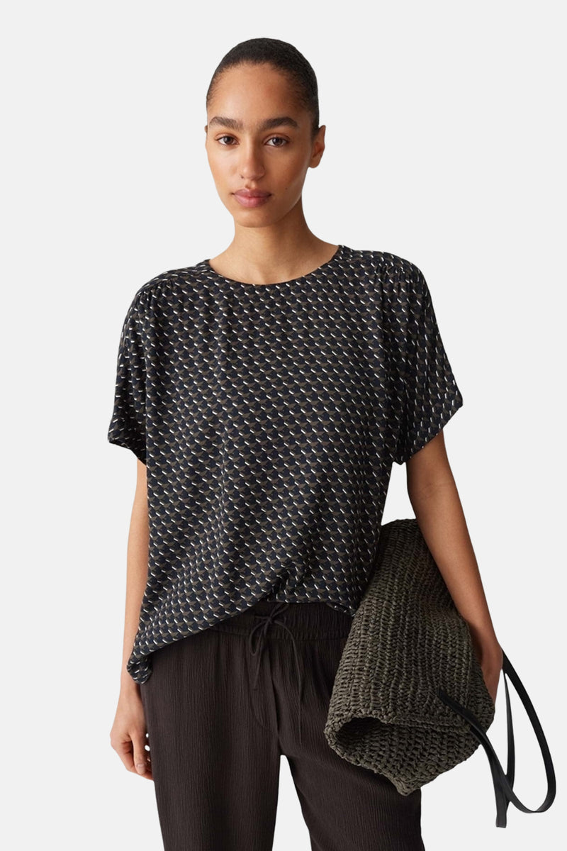 Blouse à manches courtes multicolore de OPUS, avec motif géométrique sur fond noir, portée avec un pantalon brun ample.
