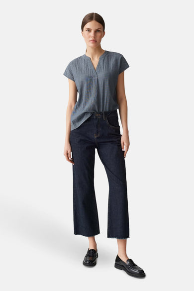 Blouse bleu à manches courtes par OPUS, avec un col en V et un motif texturé, portée avec un jean ample et des mocassins noirs.
