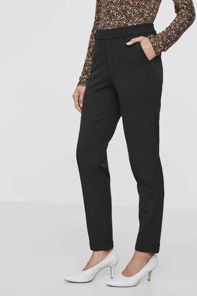 Pantalon de costume noir par VERO MODA, avec poches et revers, porté avec un chemisier à motifs floraux et des escarpins blancs.