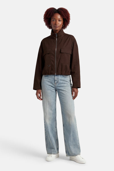Veste casual brune de OPUS avec col montant, fermeture éclair argentée et poches à rabat, portée avec un jean clair et des baskets blanches.