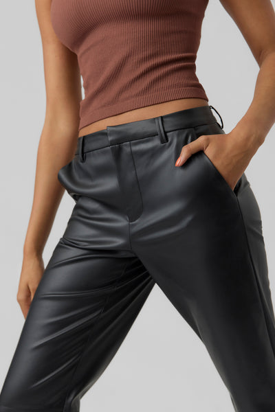 Pantalon noir en similicuir de VERO MODA, porté avec un top côtelé marron et une main dans la poche.

