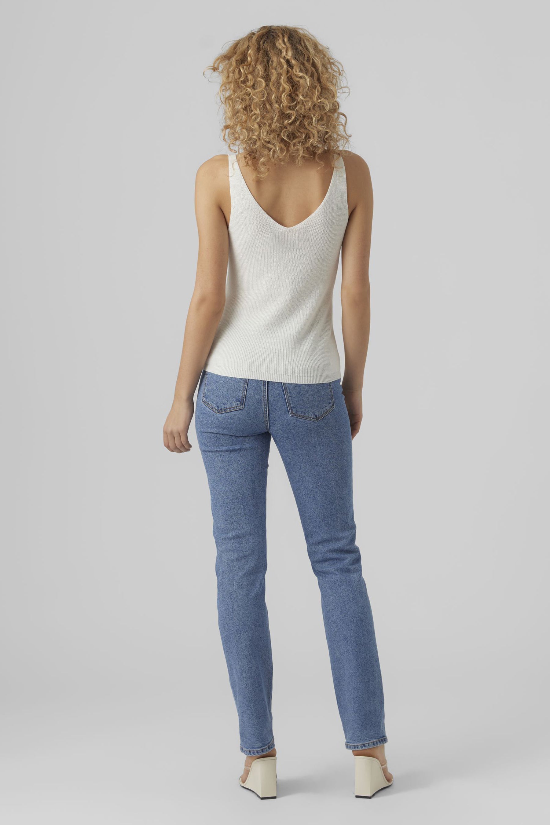 Débardeur blanc de VERO MODA, avec un dos en V, porté avec un jean bleu et des sandales à talons compensés.