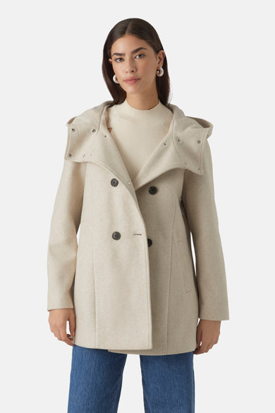 Korte beige jas van VERO MODA® met een drukknoopcapuchon, gedragen over een gebroken witte trui en een blauwe jeans.