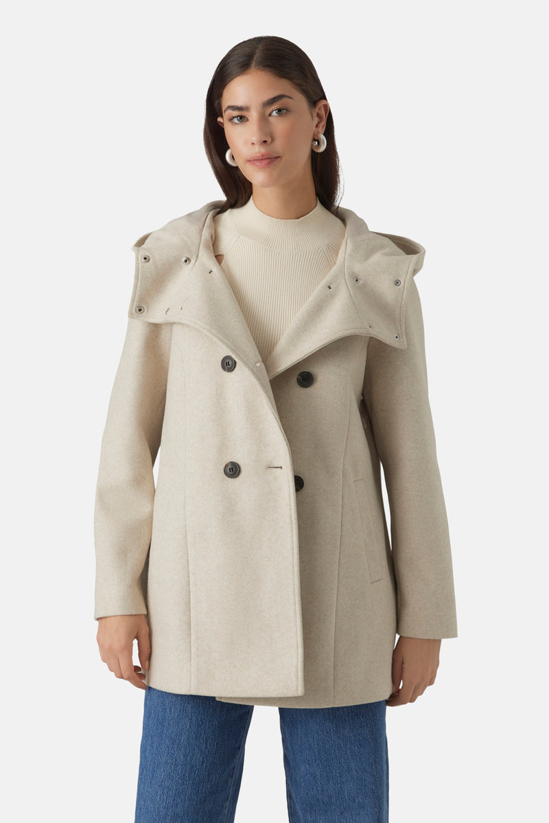 Korte beige jas van VERO MODA® met een drukknoopcapuchon, gedragen over een gebroken witte trui en een blauwe jeans.