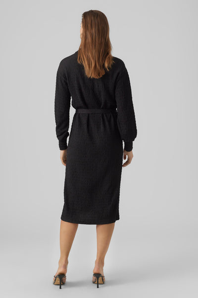 Robe midi noire par VERO MODA, à texture plissée, avec ceinture assortie et chaussures à talons noirs.