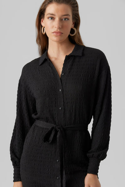 Robe midi noire de VERO MODA, avec ceinture nouée et texture froissée, portée avec des boucles d'oreilles créoles dorées.
