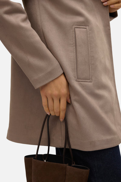 Veste longue marron de VERO MODA® avec poche passepoilée, portée sur un jean bleu marine et un sac marron.
