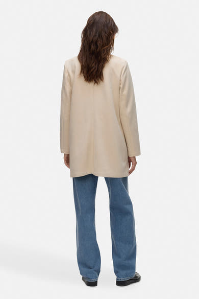 Ecru lange jas van VERO MODA, van achteren gezien, gedragen met een wijde blauwe jeans.
