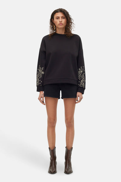 Short noir de VERO MODA, porté avec un pull noir brodé et des bottes en cuir marron.
