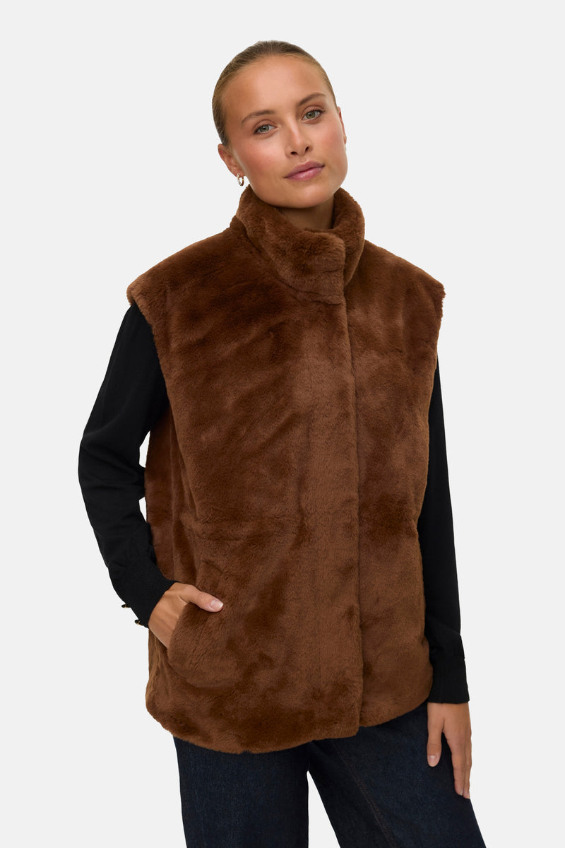 Veste sans manches marron en fausse fourrure de VERO MODA, portée sur un pull noir, avec un pantalon en denim foncé.