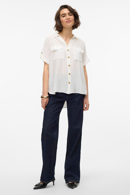 Chemisier blanc à manches courtes par VERO MODA, avec poches et boutons dorés, porté avec un jean bleu foncé.
