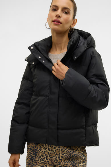 Korte zwarte jas van VERO MODA® met capuchon, gedragen over een grijze trui en een broek met luipaardprint.