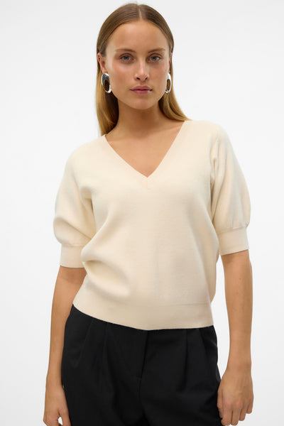 Pull à col en V écru de Vero Moda, porté avec un pantalon noir, et des boucles d'oreille argentées.
