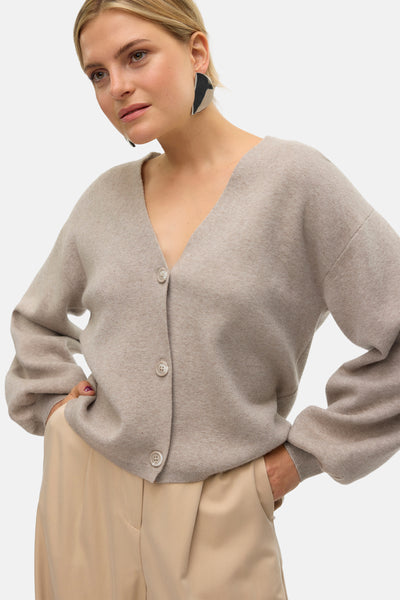 Cardigan beige de VERO MODA, boutonné sur le devant, porté avec un pantalon beige uni et des boucles d'oreilles originales.
