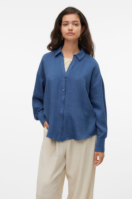 Chemisier bleu à manches longues de VERO MODA®, porté sur un débardeur en dentelle et un pantalon beige ample.
