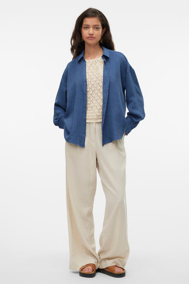 Chemise bleue à manches longues de VERO MODA, portée ouverte sur un haut en maille ajourée, avec un pantalon beige ample.

