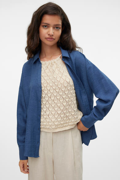 Chemise bleue à manches longues de VERO MODA®, portée ouverte sur un haut texturé et un pantalon beige.
