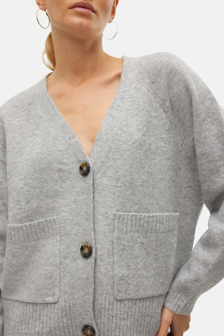Cardigan gris de VERO MODA avec poches plaquées et fermeture boutonnée marron, porté avec des boucles d'oreilles dorées.
