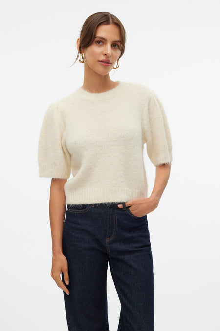 Pull à col rond écru de VERO MODA, porté avec un jean foncé et des boucles d'oreilles dorées.

