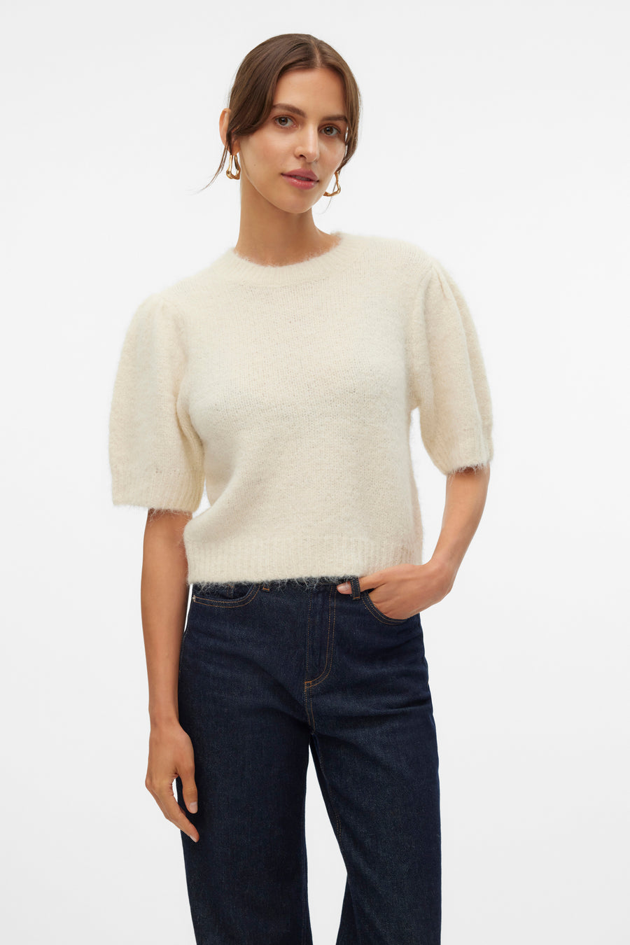 Pull à col rond écru de VERO MODA, porté avec un jean foncé et des boucles d'oreilles dorées.
