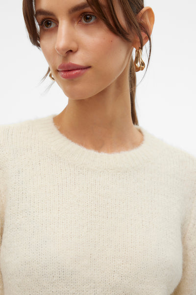 Pull à col rond écru de VERO MODA®, texture tricotée douce, porté avec des boucles d'oreilles dorées.
