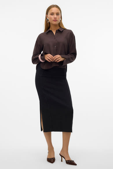 Jupe longue en denim noir de VERO MODA, avec une fente latérale et portée avec un chemisier marron et des mules.
