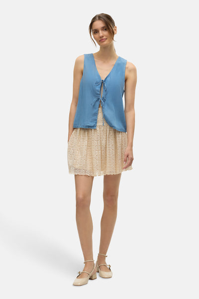 Blouse bleue de VERO MODA, avec fermeture nouée, portée sur une robe en dentelle écrue et chaussures à bride.
