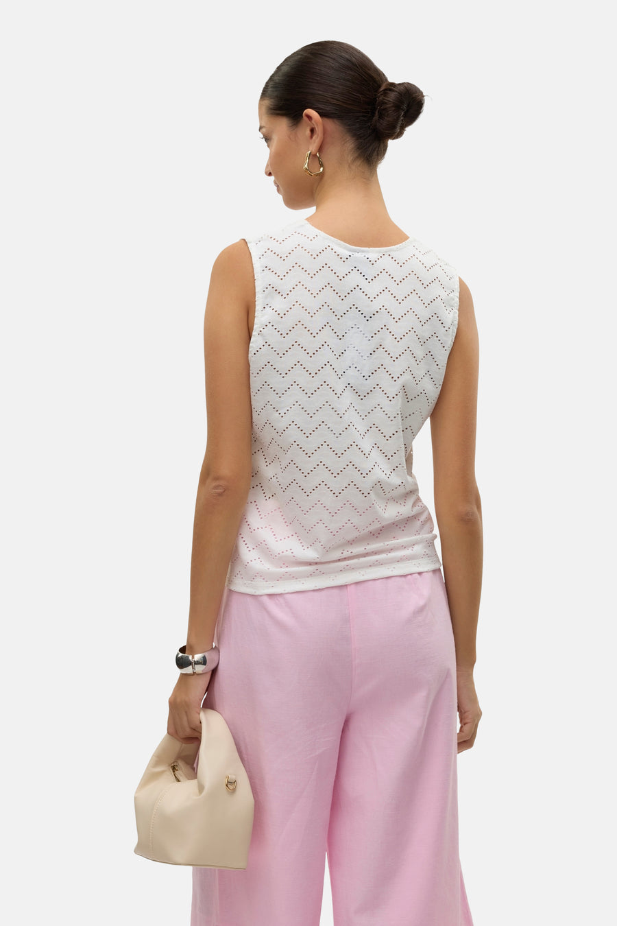 Blouse sans manches blanche ajourée de Vero Moda, avec motif à chevrons, portée avec un pantalon rose et des bijoux dorés.