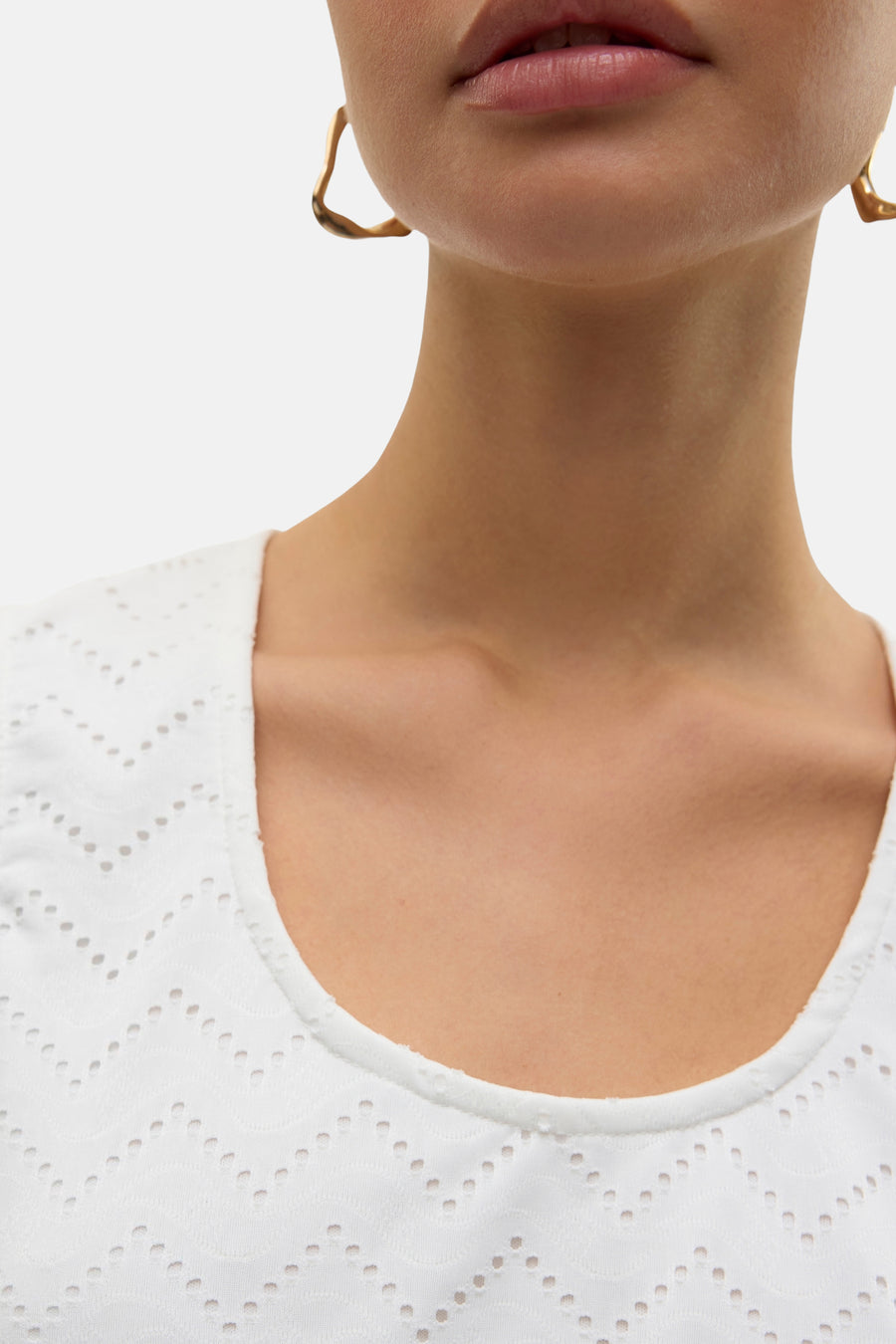 Blouse blanche sans manches de VERO MODA, en broderie anglaise, portée avec des boucles d'oreilles dorées.