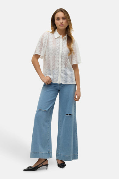 Chemisier à manches courtes blanc VERO MODA®, en dentelle florale, porté avec un jean ample et des chaussures noires.
