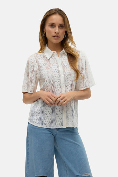 Chemisier blanc en dentelle de VERO MODA, avec des détails ajourés et porté avec un jean bleu.
