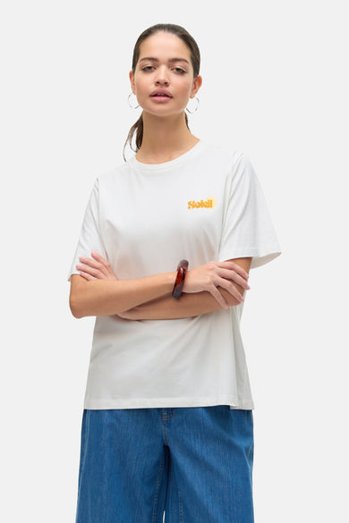 T-shirt blanc à manches courtes de Vero Moda, orné d'un motif Soleil sur le devant, porté avec un bracelet marron et un pantalon bleu.