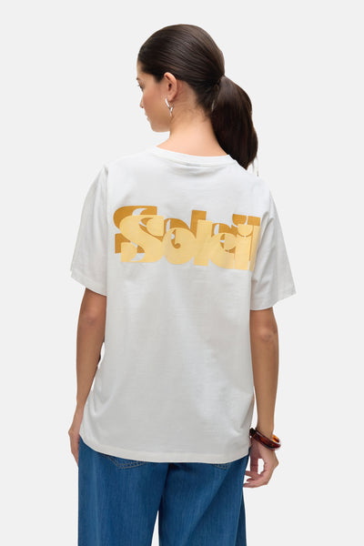 T-shirt blanc à manches courtes de VERO MODA, avec un imprimé doré dans le dos, porté avec un jean bleu.
