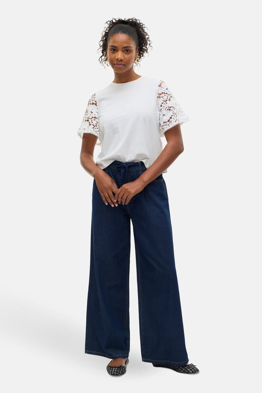 Jean large en denim bleu foncé de Vero Moda, porté avec un T-shirt blanc à manches en dentelle et des ballerines à carreaux.
