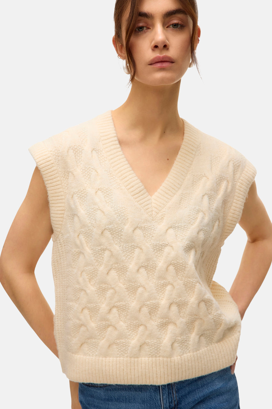 Débardeur beige à torsades par VERO MODA, porté avec un jean bleu, modèle avec les mains sur les hanches.
