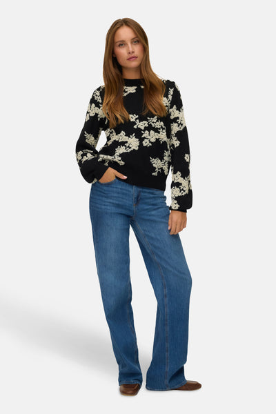 Pull à col rond noir de Vero Moda, à motif floral blanc, porté avec un jean bleu et des chaussures marron.