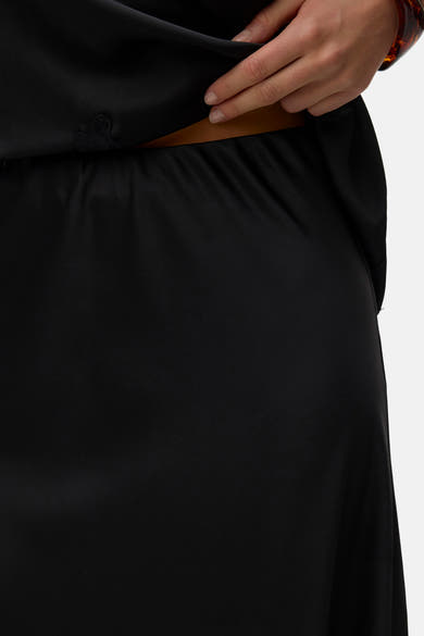 Black long skirt with smooth satin texture and subtle side seam detail, cropped view showing waistband and hand adjusting fabric