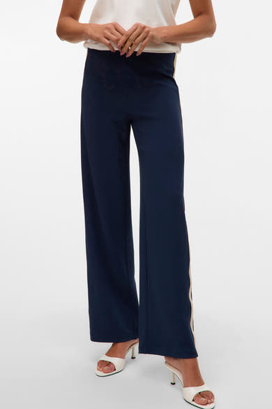 Pantalon bleu marine à coupe droite, par VERO MODA®, avec bande latérale beige et chaussures blanches à talons.
