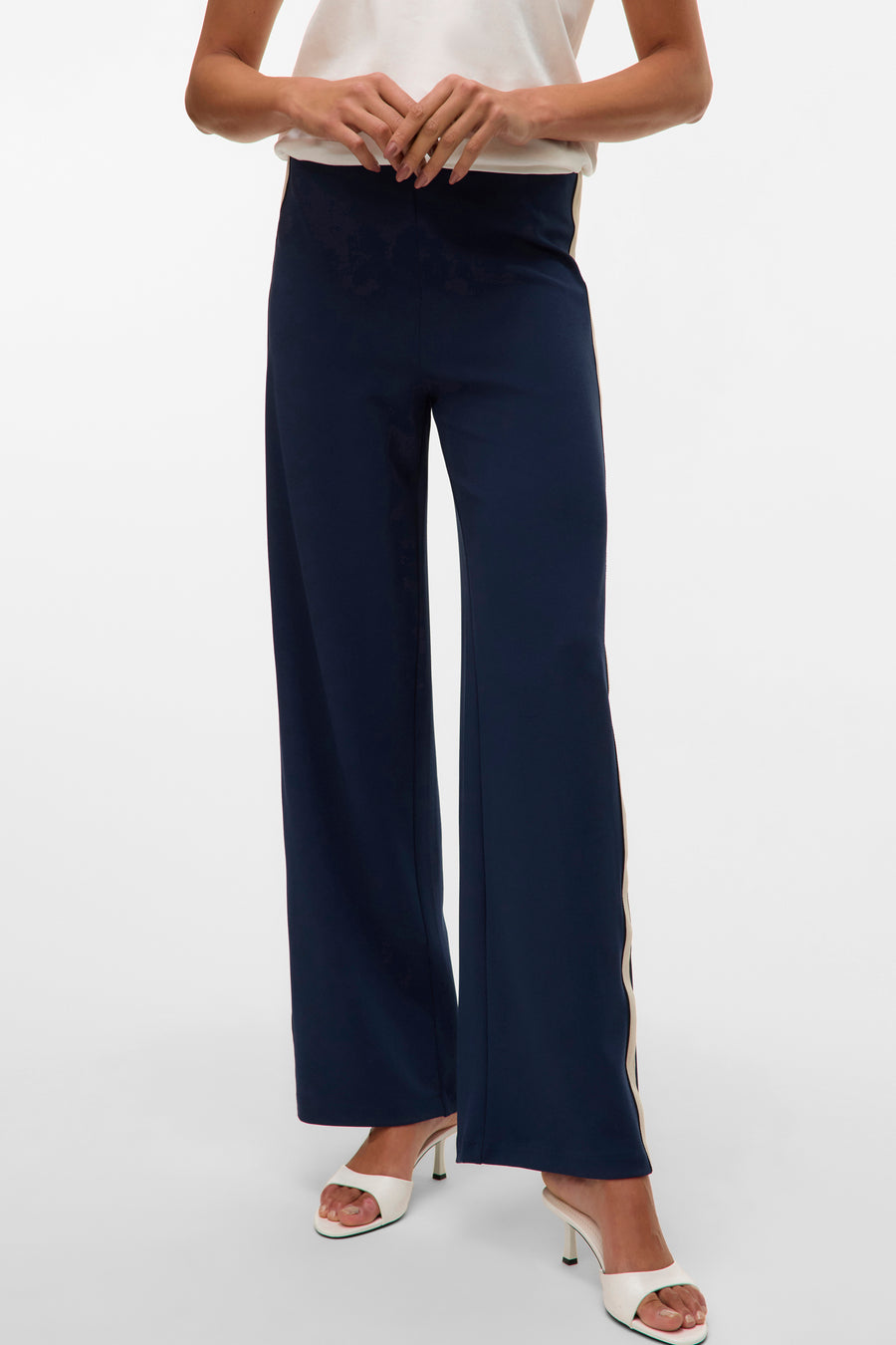 Pantalon bleu marine à coupe droite, par VERO MODA®, avec bande latérale beige et chaussures blanches à talons.
