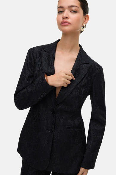 Zwarte blazer van VERO MODA, getextureerd met bloemenpatroon, open gedragen met gouden oorbellen.