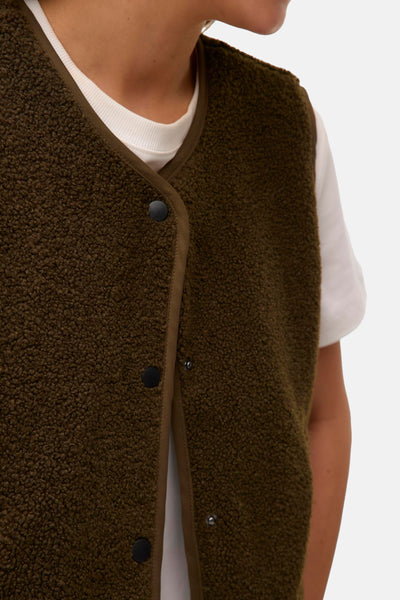 Groen schapenvachtimitatie gilet van VERO MODA, camelkleurige bies en zwarte drukknoppen, gedragen over een wit T-shirt.