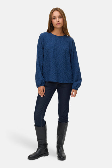 Chemisier bleu à manches longues par VERO MODA, avec une texture gaufrée, porté avec un jean foncé et des bottes noires.
