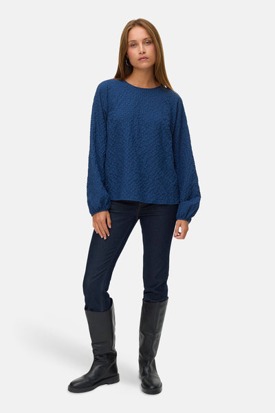 Chemisier bleu à manches longues par VERO MODA, avec une texture gaufrée, porté avec un jean foncé et des bottes noires.

