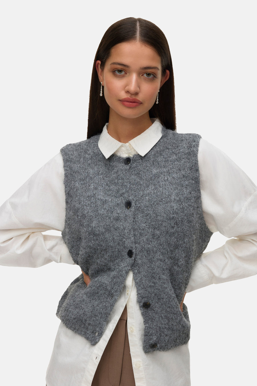 Débardeur gris en tricot par Vero Moda, porté sur une chemise blanche à manches longues avec des boucles d'oreilles pendantes.